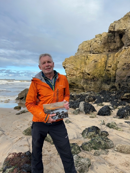 Man standing on beach in front of rock formation holding book