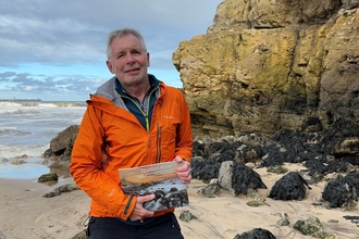 Man standing on beach in front of rock formation holding book