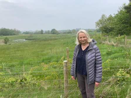 Woman in grey coat standing in front of fence with grass and stream area behind