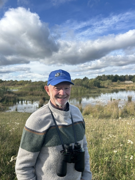 man wearing cap and binoculars standing in front of grasslands and  pond