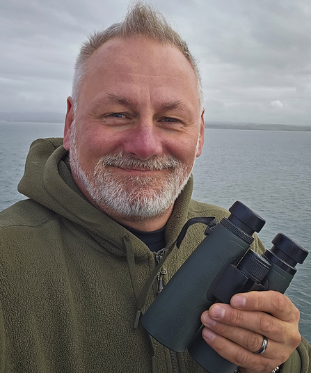 Trustee Ian Cole in front of water holding binoculars